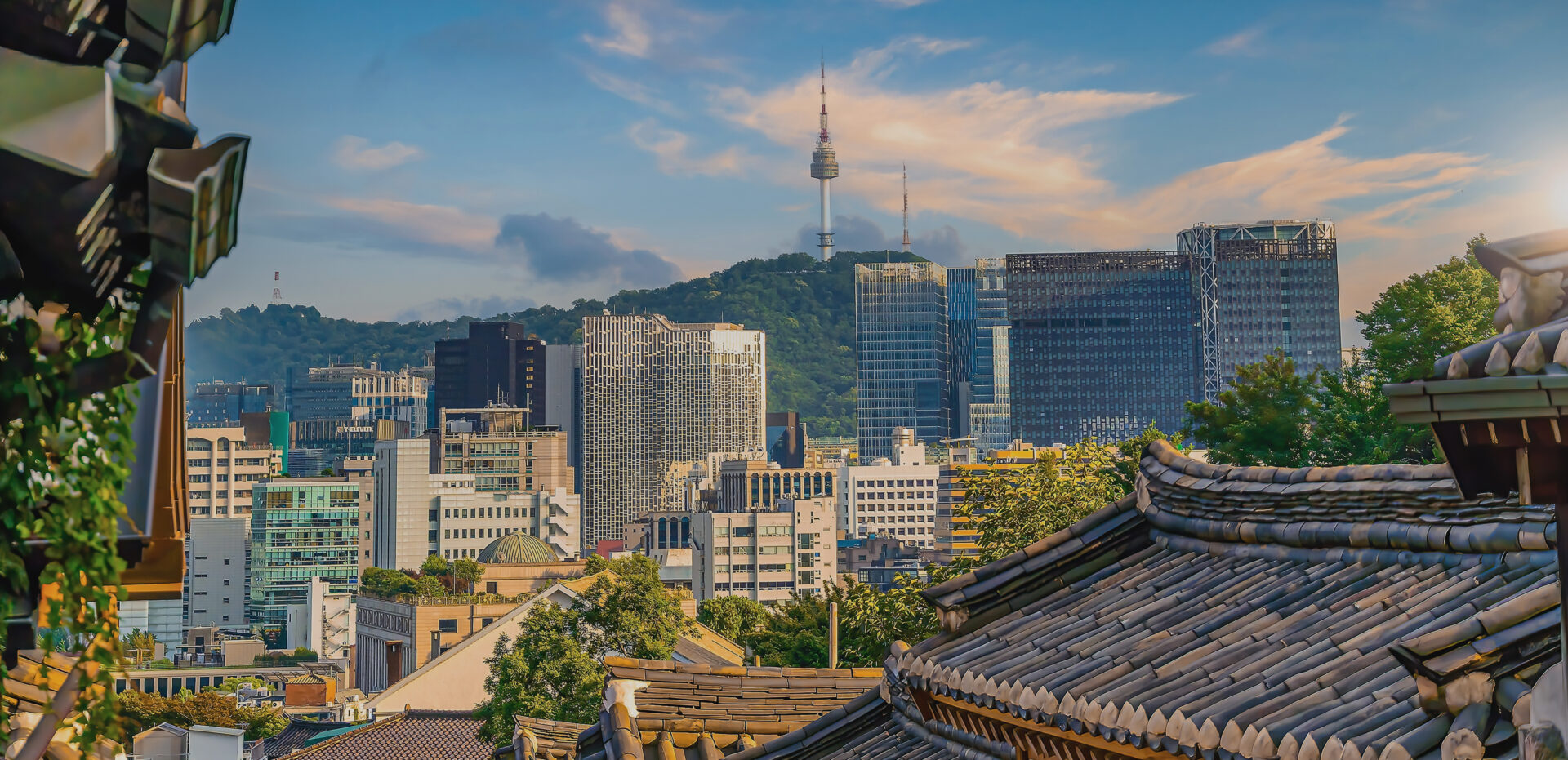 City skyline with modern glass buildings behind terracotta-tiled rooftops and a green hill in the distance.