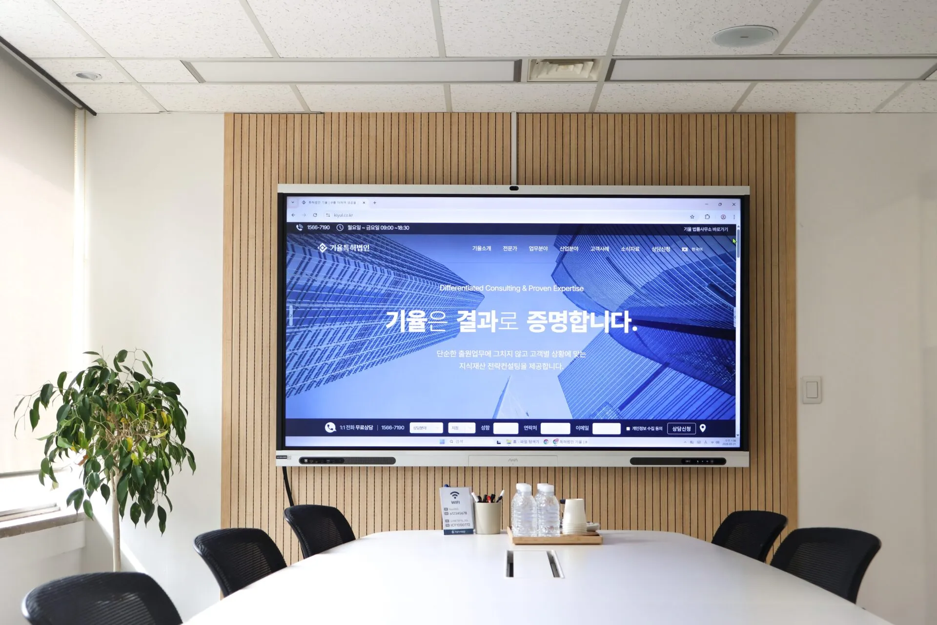 Conference room with a large wall-mounted screen showing a blue website, wooden slat wall, and plant in the corner. Attention: wall-mounted display behind a meeting table with water bottles and cups nearby.